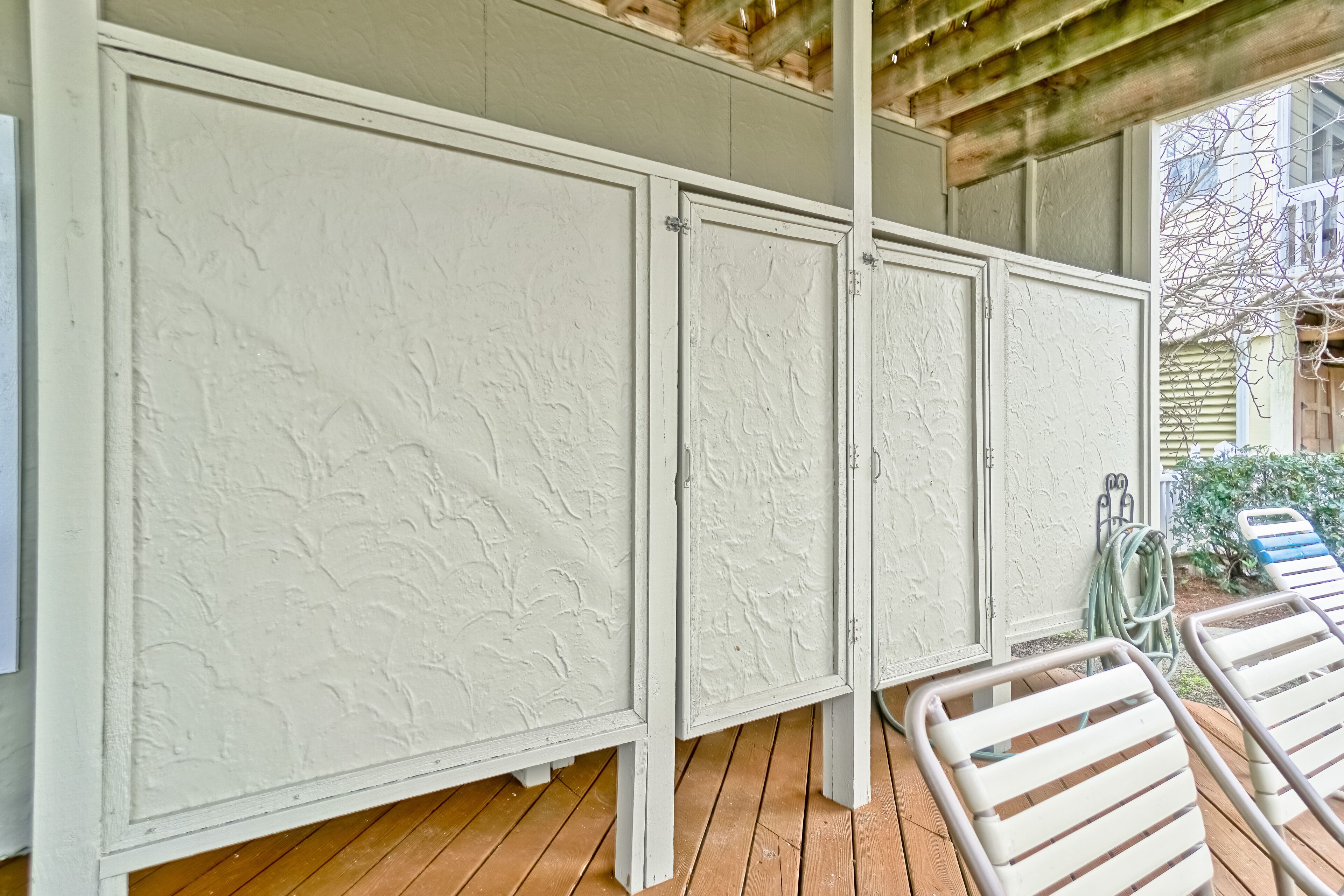 A covered outdoor area with a wooden deck, white textured walls, and metal chairs.
