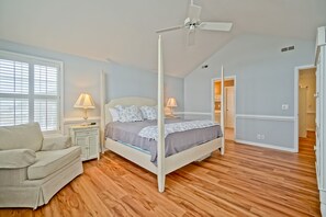 Een slaapkamer met een hemelbed, houten vloeren, een witte plafondventilator en een raam met zonwering.