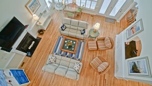 A spacious living room with a large sofa, a coffee table, and a TV. There are multiple windows providing natural light and a view of the outdoors.