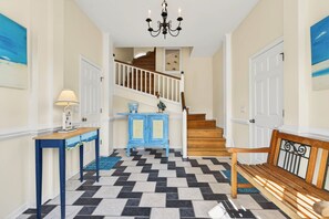 A hallway with a checkered floor, a staircase, a blue cabinet, a wooden bench, and a chandelier.