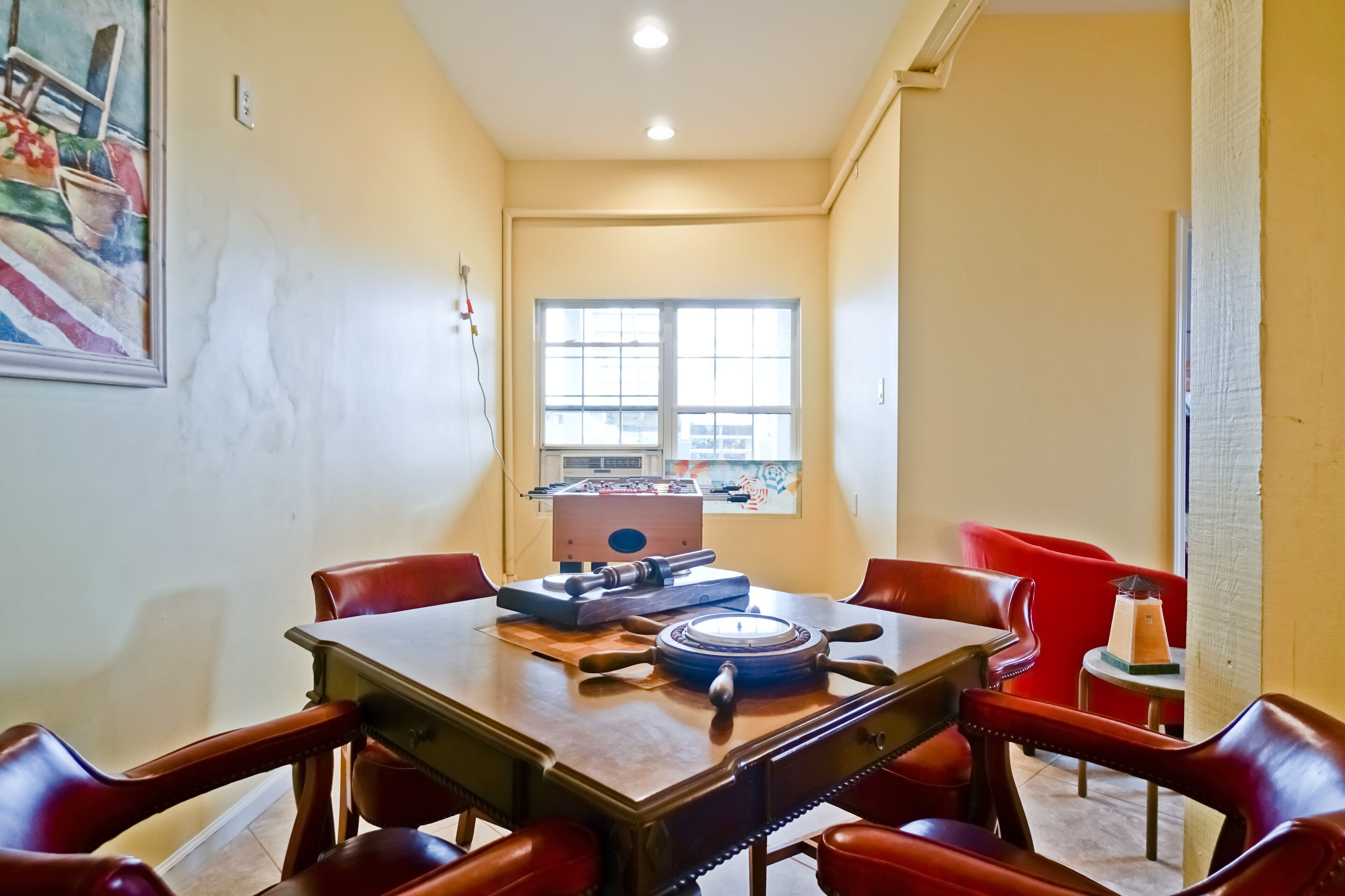A dining area with a wooden table and red chairs, a window with a view, and a painting on the wall.