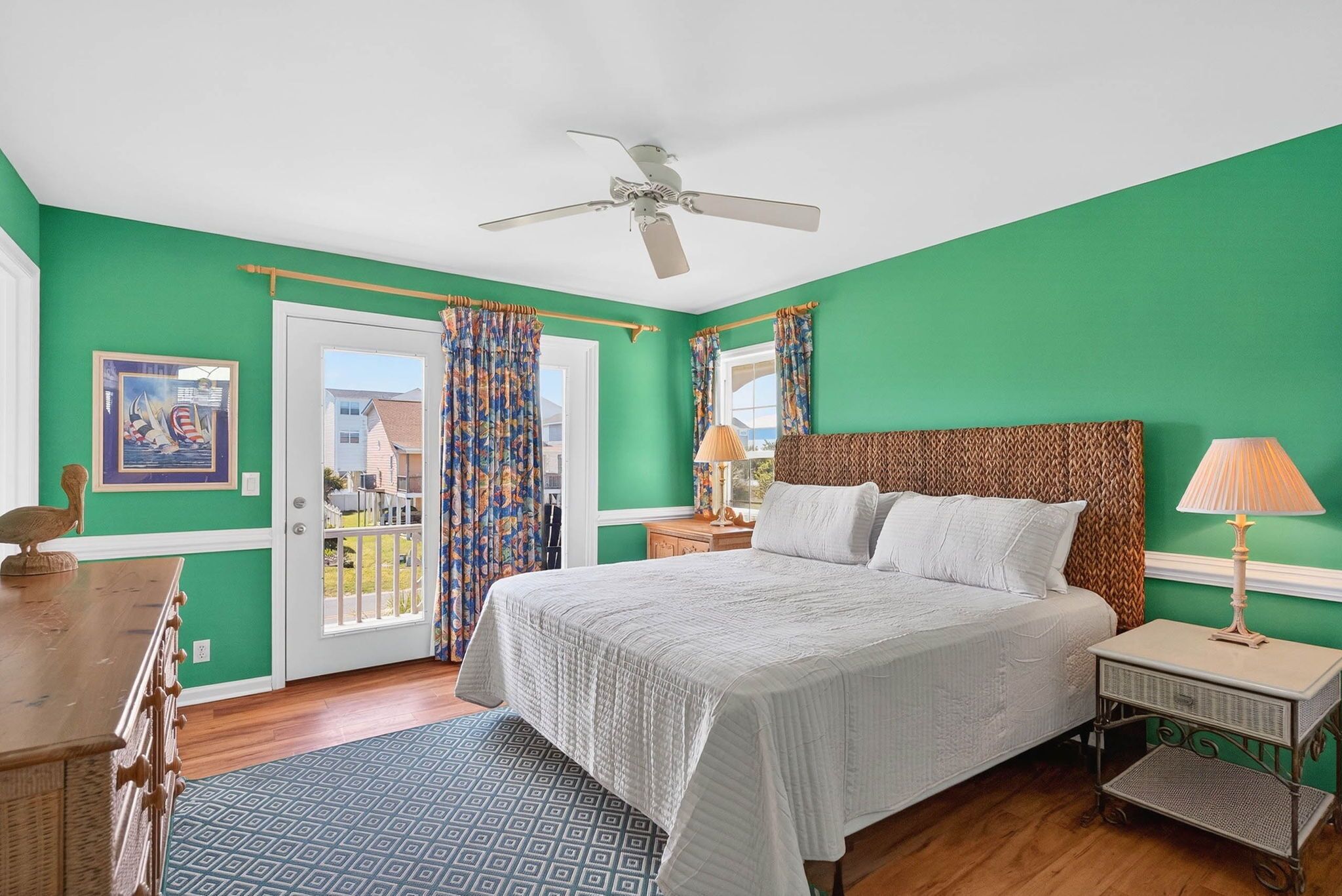 Un dormitorio con una cama, una mesita de noche, una lámpara, un ventilador de techo, una ventana con cortinas y un cuadro en la pared.
