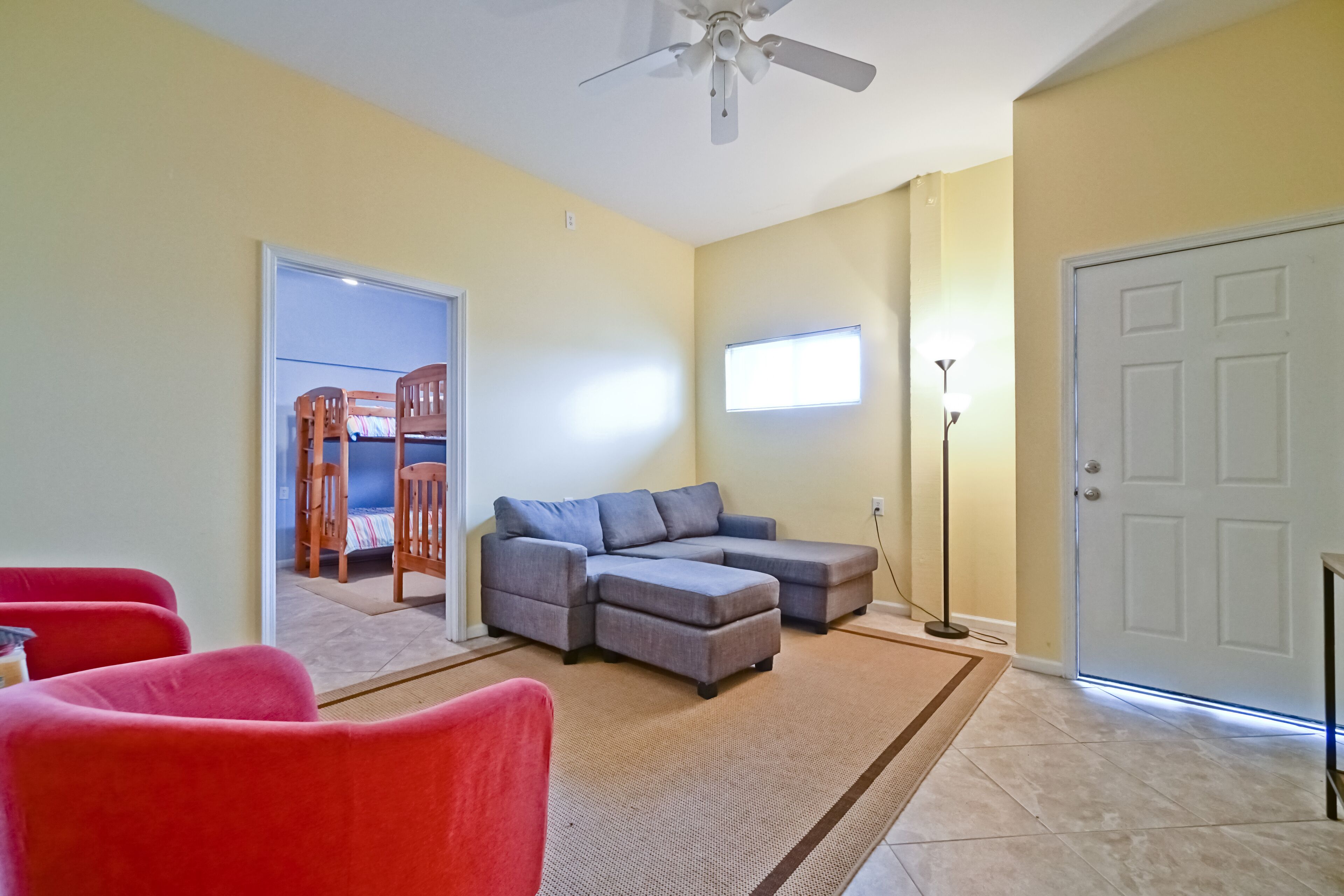A living room with a sectional sofa, red chairs, a ceiling fan, and a door.