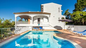 A swimming pool with lounge chairs and a covered seating area, surrounded by a white building with arches and a tiled roof.