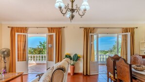 A spacious living room with a sofa, dining table, and chairs, a fan, and a chandelier. There are open doors leading to a balcony with a view of greenery.