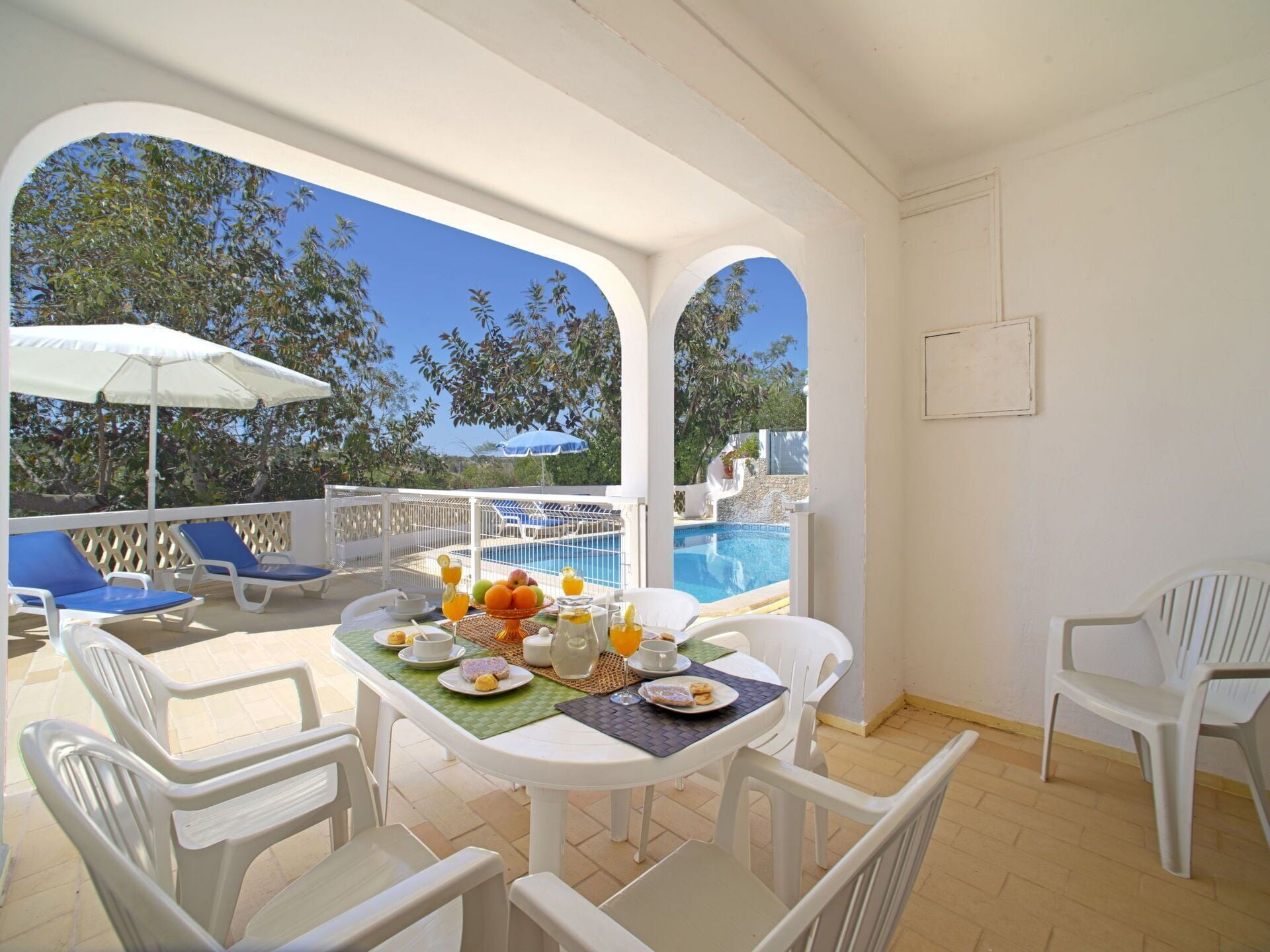 A sunlit outdoor dining area with a pool view, white furniture, and a table set with fruits and beverages.