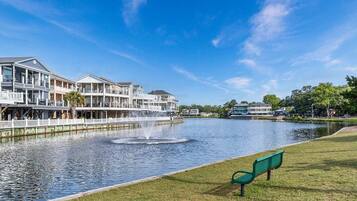 A waterfront property with a fountain, a grassy area with a bench, and a row of multi-story buildings.