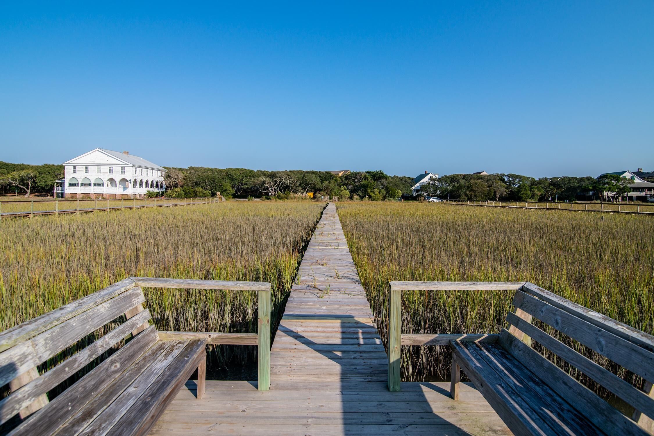 A wooden boardwalk leading to a white building with arched windows, surrounded by a field.