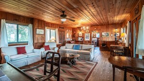 A cozy cabin interior with wooden walls and ceilings, furnished with two white sofas, a coffee table, and a dining table. - Ocean Breezes Historic Home with Creek Dock (Pawleys Island)