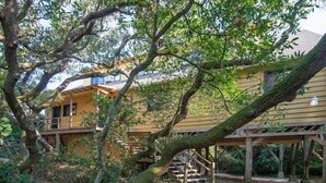 A rustic wooden cabin with a wooden deck and staircase, surrounded by dense foliage. - Ocean Breezes Historic Home with Creek Dock (Pawleys Island)