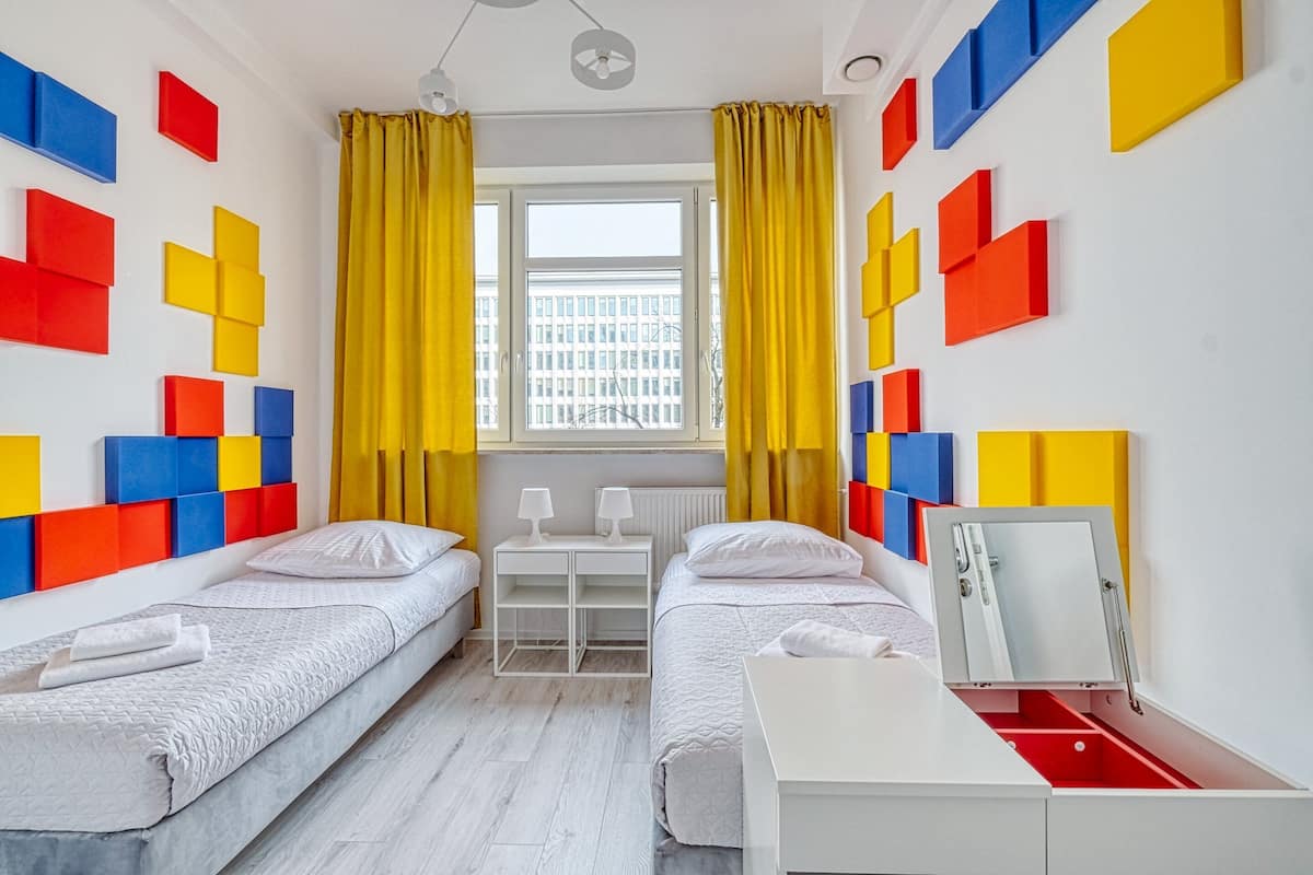 Quarto com duas camas, uma escrivaninha e uma janela com cortinas amarelas.