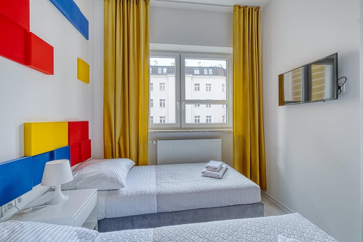 Quarto de hotel com duas camas, uma janela com cortinas amarelas, uma TV fixada na parede e um radiador.
