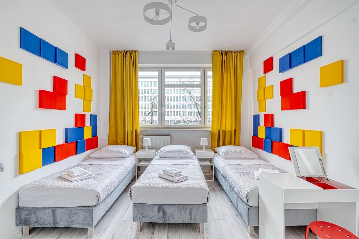 Quarto moderno com duas camas, uma escrivaninha e vista para um edifício através de grandes janelas.