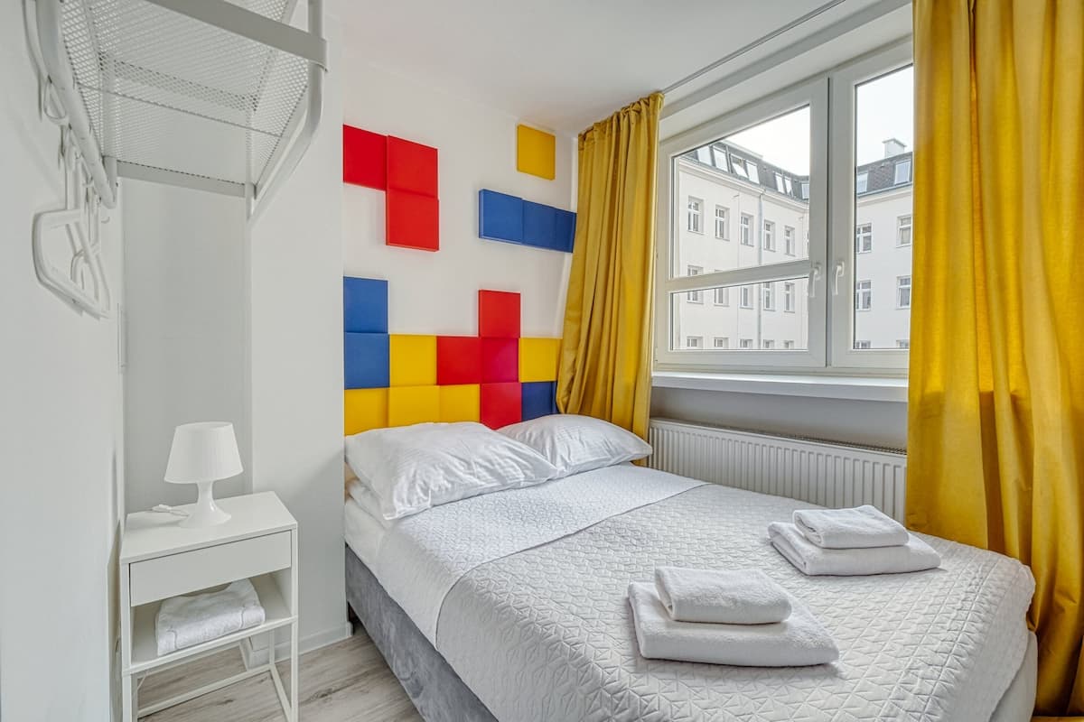 Quarto de hotel com cama, mesa de cabeceira, janela com cortinas e parede com padrões geométricos coloridos.