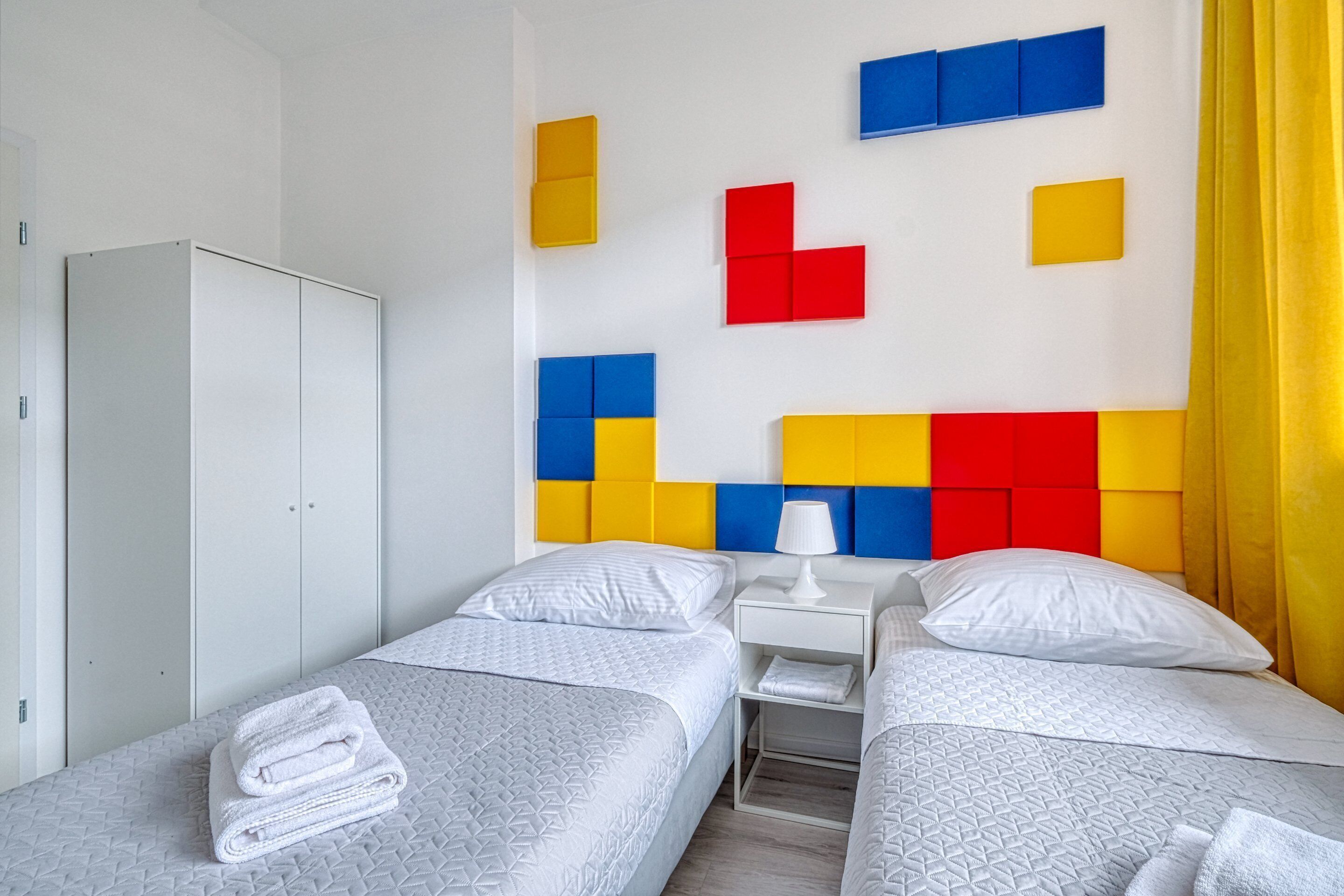 A hotel room with two beds, a wardrobe, and a wall with colorful geometric panels.