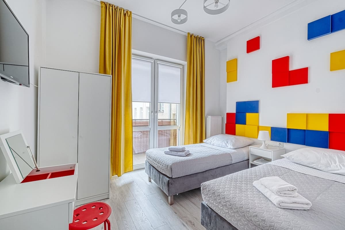 Quarto de hotel moderno com duas camas, um guarda-roupa, uma escrivaninha e uma janela com cortinas amarelas.