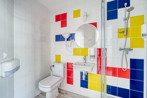 Une salle de bain comprenant des toilettes, un lavabo et une douche. Les murs sont recouverts de carreaux aux couleurs rouge, jaune et bleu, disposés en motifs géométriques.
