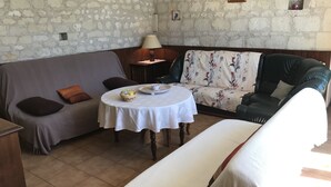 Une pièce chaleureuse avec un mur en pierre, un canapé gris, un fauteuil à motifs floraux, une table ronde recouverte d’une nappe blanche et une petite lampe.