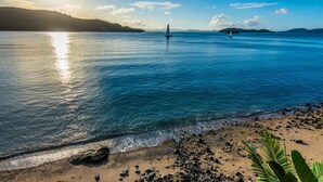 Une plage à l’eau claire, un voilier et des collines qui se dessinent à l’horizon.