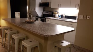 A kitchen with a granite countertop, white bar stools, a microwave, and a sink. - Ocean‑View at Island Surf- Walk to Beaches, Surfing, Shops, Dining & Kalama Park (Kihei)