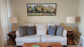 A living room with a light-colored sofa, patterned pillows, two wooden side tables with lamps, and a painting of a tropical landscape above the sofa.