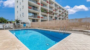 A swimming pool with a clear blue water, surrounded by a tiled deck. In the background, there is a multi-story building with balconies, some with plants. - Ocean‑View at Island Surf- Walk to Beaches, Surfing, Shops, Dining & Kalama Park (Kihei)