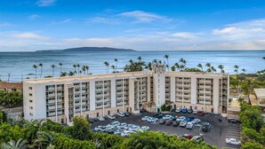 A multi-story hotel with a parking lot and a view of the ocean. - Ocean‑View at Island Surf- Walk to Beaches, Surfing, Shops, Dining & Kalama Park (Kihei)