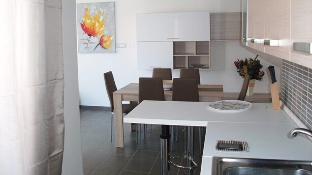 A modern kitchen with a white countertop, a sink, and a dining area with a white table and brown chairs. - Bella casa vacanze in Sardegna (Lotzorai)