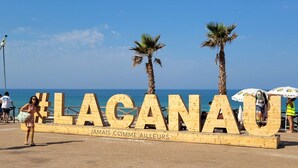 Une pancarte en bois portant les inscriptions « LAGANAU » et « JAMAIS COMME AILLEURS », placée sur une plage bordée de palmiers, avec la mer en arrière-plan.