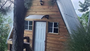 Una cabaña de madera con puerta blanca, un ventanal pequeño y una escalera que lleva a la entrada.