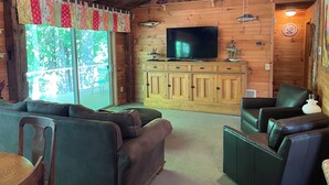 A cozy cabin interior with a wooden ceiling, a large TV on a wooden cabinet, and a sliding glass door leading to an outdoor deck. - Beautiful Cabin with Gorgeous Riverbend View. Great for relaxation and fishing! (Branch Township)