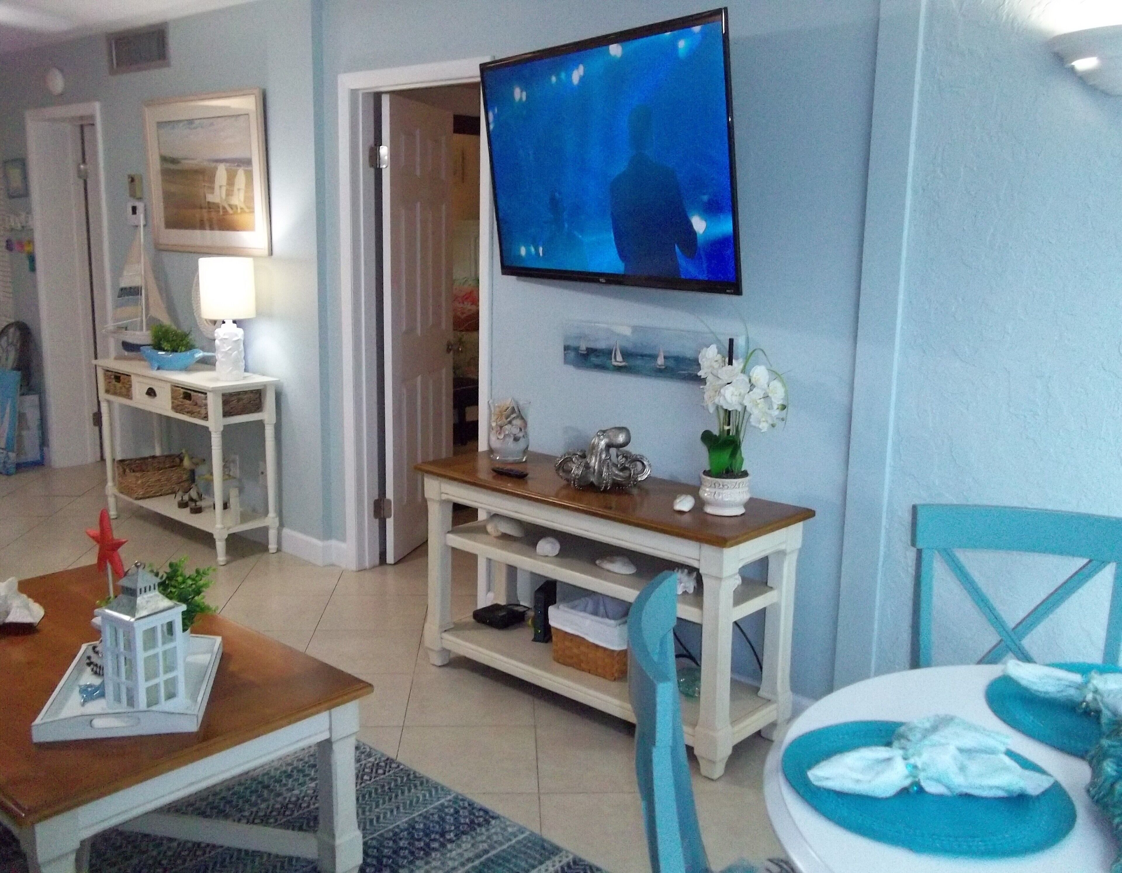 A living room with a TV mounted on the wall, a wooden coffee table, a dining table with chairs, and a small decorative plant.