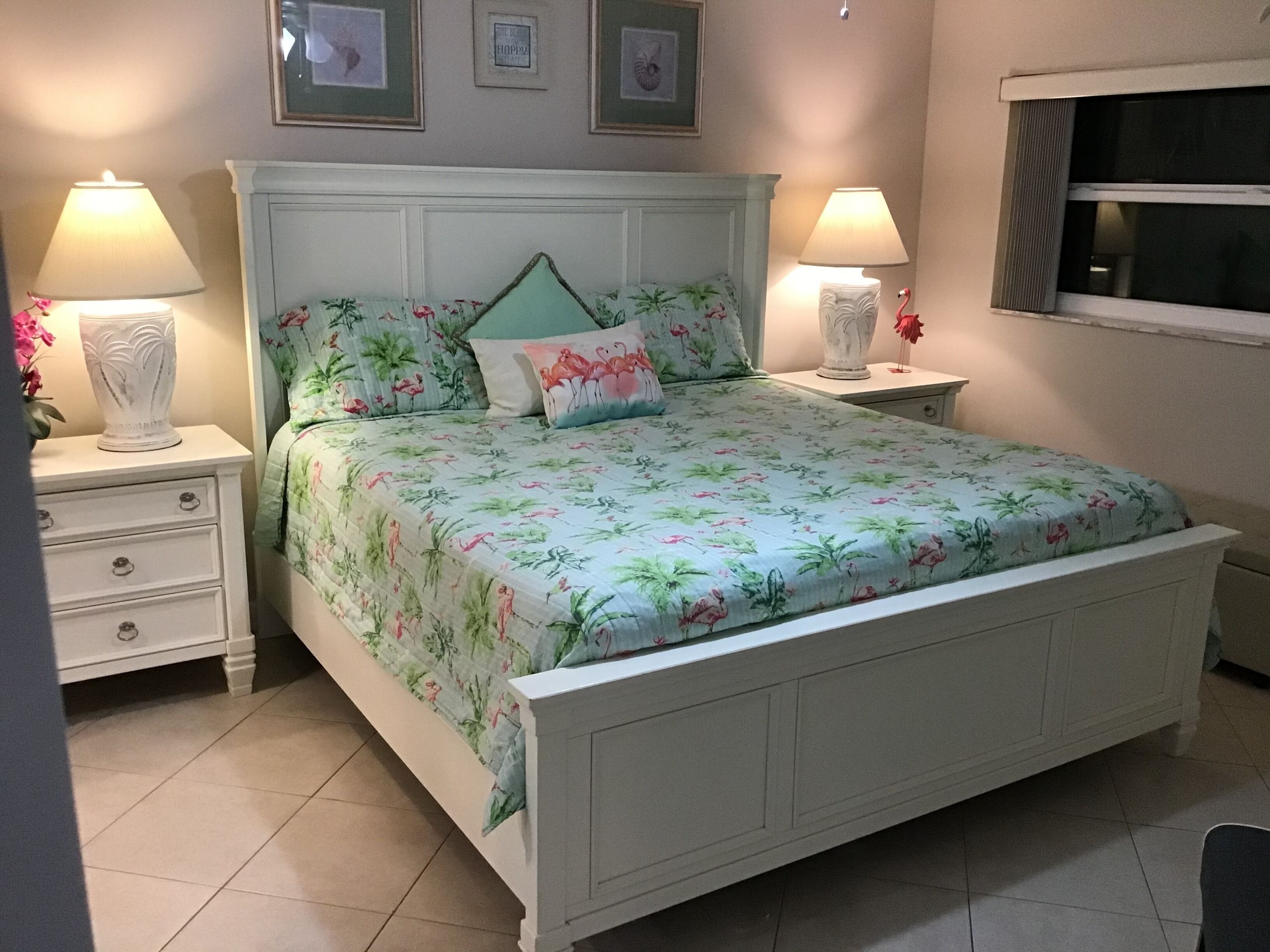 Een slaapkamer met een bloemenbedekking, twee bedlampjes, een nachtkastje met lades en een raam met jaloezieën.