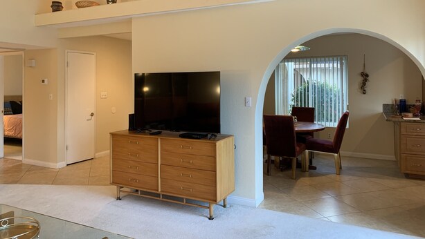 A spacious living area with a TV, wooden cabinet, dining table, and arched doorway. - Tranquil 3BR, 2BA Condo at Indian Palms - near Coachella (Indio)