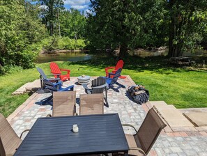 Een terras buiten met een tafel en stoelen, een vuurkuil en uitzicht op een vijver omgeven door bomen.