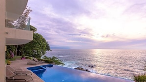A modern infinity pool with a view of the ocean, lounge chairs, and a building with a glass balcony. - Exclusive Penthouse Oceanfront, small Condominium complex (Conchas Chinas)