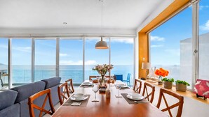 A dining area with a wooden table set for six, large windows offering a view of the sea, and a sofa with cushions. - Exclusive Penthouse Oceanfront, small Condominium complex (Conchas Chinas)