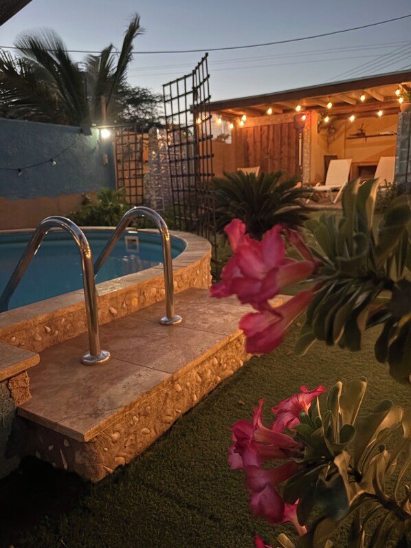A swimming pool with a metal ladder, surrounded by artificial grass and tropical plants.