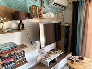 A modern living room with a flat-screen TV mounted on a wooden wall, a bookshelf with various books, decorative items, and a small table with a lamp and remote controls.