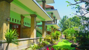 Un edificio de varios pisos con una combinación de colores verde y beige, que cuenta con un jardín con diversas plantas y un cielo despejado.