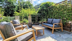An outdoor patio with wooden furniture, including chairs and a sofa, surrounded by lush greenery and a wooden trellis.