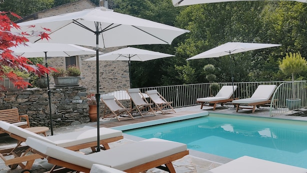 A poolside area with lounge chairs and umbrellas, a stone wall, and a view of a house in the background.