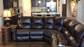A modern living room with a large L-shaped sectional sofa, a kitchen with dark cabinetry, and a dining area with a white table and chairs. - Lakefront home with private beach  & unbelievable sunsets on Lake Keowee (Seneca)