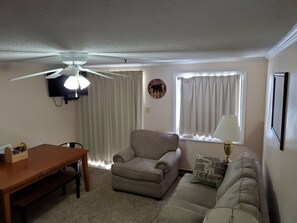 Una sala de estar con ventilador de techo, un sofá, una silla, una mesa y una ventana con cortinas.