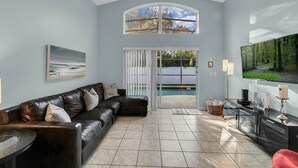 A modern living room with a leather sofa, a flat-screen TV, and a large window overlooking a pool. - Disney home- Private Pool&Hot Tub! 4b/3ba, 7 beds!! Close to all the  parks!!! (Kissimmee)