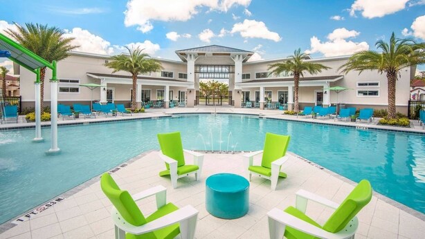 A pool area with a central seating area, palm trees, and a building with multiple windows in the background. - Disney home- Private Pool&Hot Tub! 4b/3ba, 7 beds!! Close to all the  parks!!! (Kissimmee)
