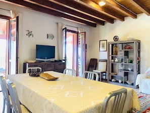 A dining area with a table set for four, a television on a wooden cabinet, and a bookshelf with various items.