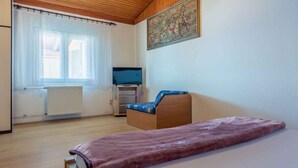 Un dormitorio con cama, una silla de madera, un televisor sobre un soporte, un radiador y una ventana con cortinas.