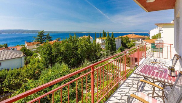 En balkon med udsigt over havet, omgivet af grøn vegetation og bygninger.