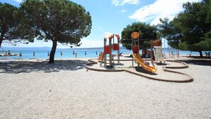 A playground with slides and climbing structures, surrounded by trees and a beach with umbrellas. - Holiday apartment with balcony and air conditioning (Crikvenica)